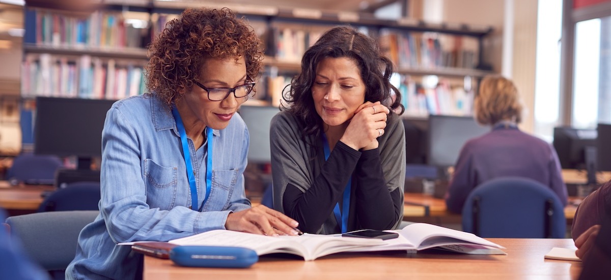 Teacher With Mature Female Adult Student Sitting At Table Working In College Library