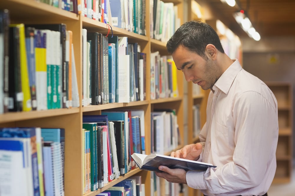 Intellectual man reading a book standing in library in front of bookshelves