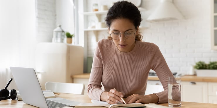 Woman learning at home referencing a book while on her laptop. 