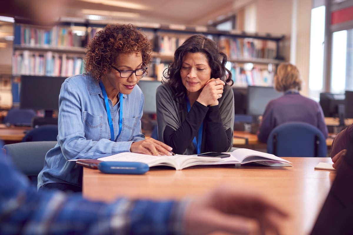 Teacher With Mature Female Adult Student Sitting At Table Working In College Library